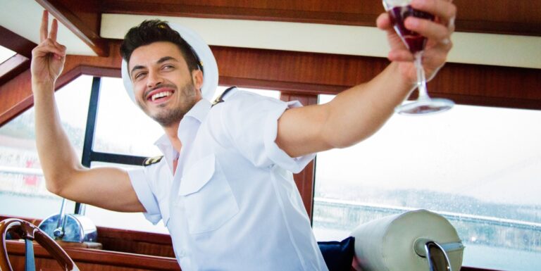 Image of a man posing with a glass of wine while sitting behind the steering wheel of a boat