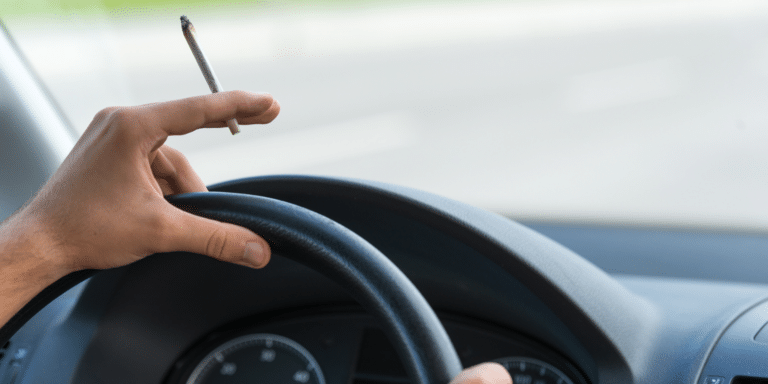 Image of a someone driving a car, and holding a marijuana cigarette in one hand.